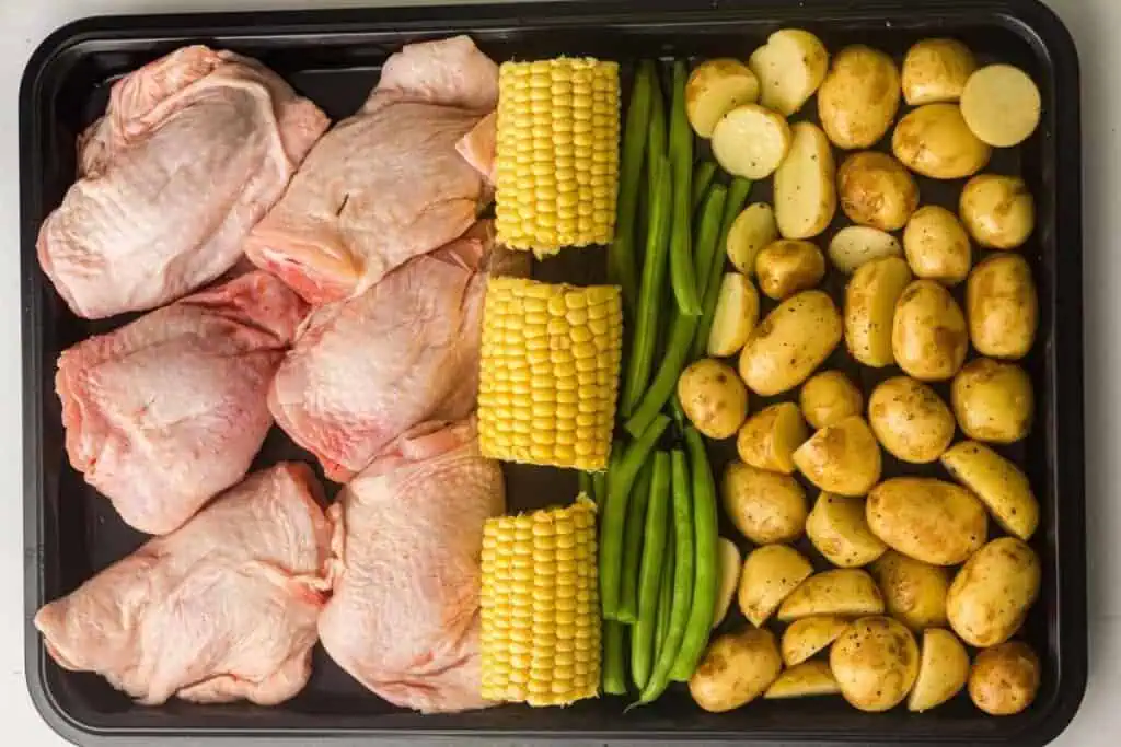 A baking sheet with raw chicken thighs, corn on the cob, green beans, and halved baby potatoes arranged in rows.