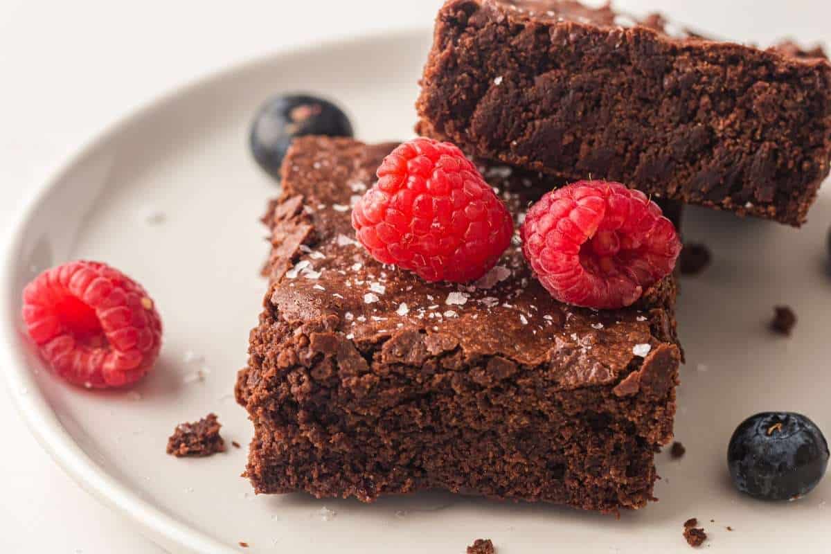 Two Sheet pan chocolate brownies on a white plate, topped with raspberries and a sprinkle of sea salt, with additional raspberries and blueberries on the side.