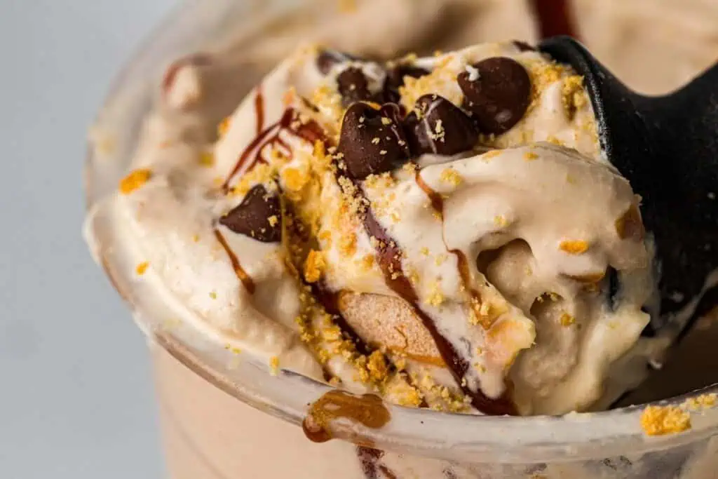 Close-up of a creamy dessert in a plastic cup, topped with chocolate chips, cookie crumbs, and a drizzle of chocolate syrup.