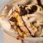 Close-up of a creamy dessert in a plastic cup, topped with chocolate chips, cookie crumbs, and a drizzle of chocolate syrup.