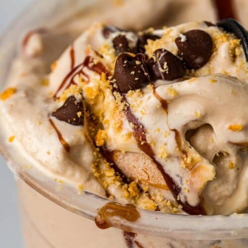 Close-up of a creamy dessert in a plastic cup, topped with chocolate chips, cookie crumbs, and a drizzle of chocolate syrup.