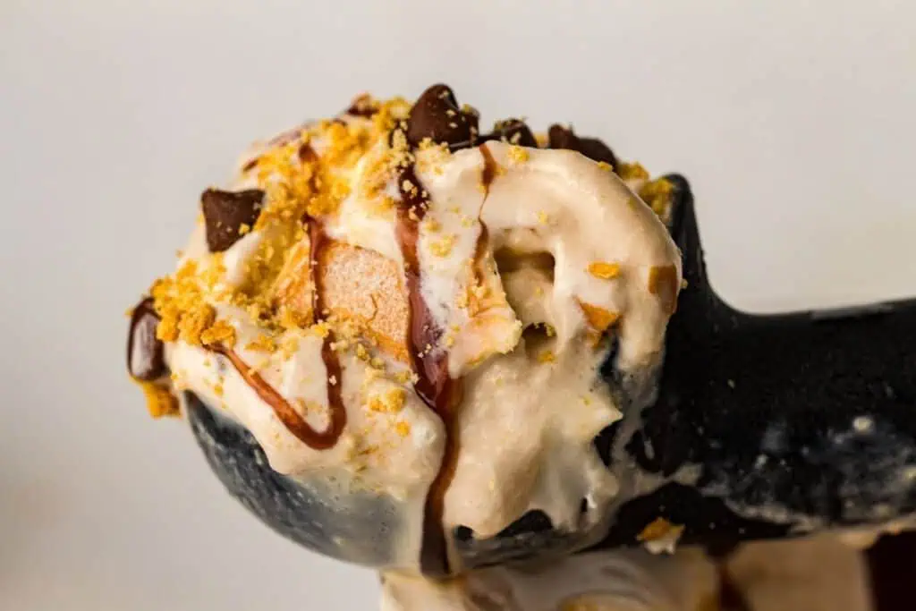 A close-up of an ice cream scoop topped with chocolate chips, crushed cookies, and a drizzle of chocolate sauce.