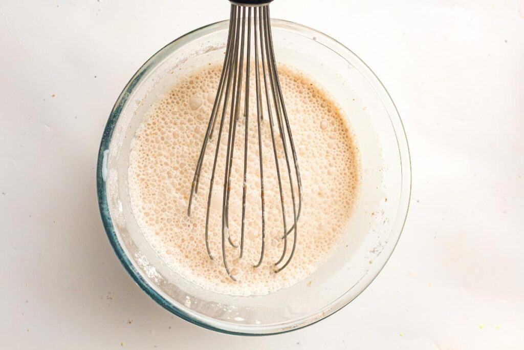 A metal whisk in a glass bowl mixing a bubbly beige liquid batter on a white surface.