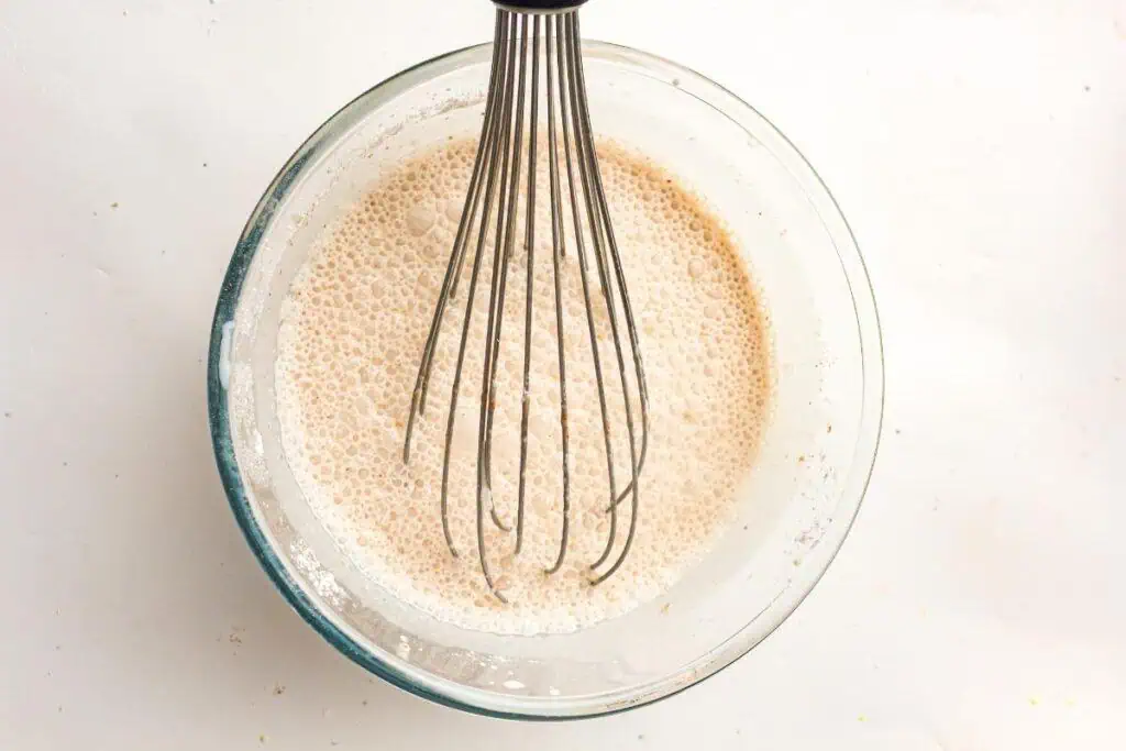 A metal whisk in a glass bowl mixing a bubbly beige liquid batter on a white surface.