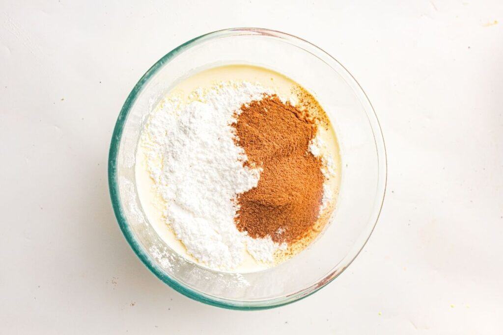 A glass bowl containing a mixture of white powdered sugar, brown cinnamon, and a creamy liquid on a white surface.