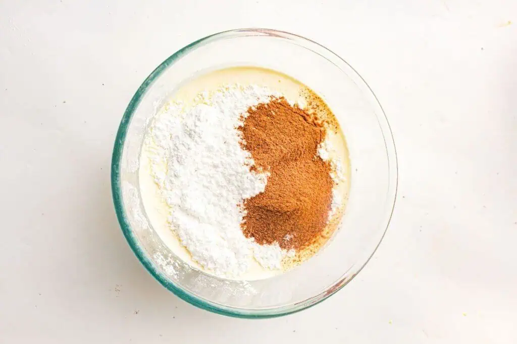 A glass bowl containing a mixture of white powdered sugar, brown cinnamon, and a creamy liquid on a white surface.