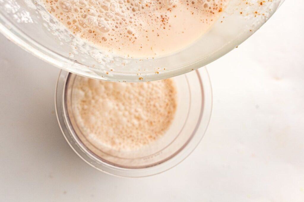 A glass bowl with a frothy, light brown liquid is being poured into a clear cup containing a similar foamy beverage, seen from above on a white surface.