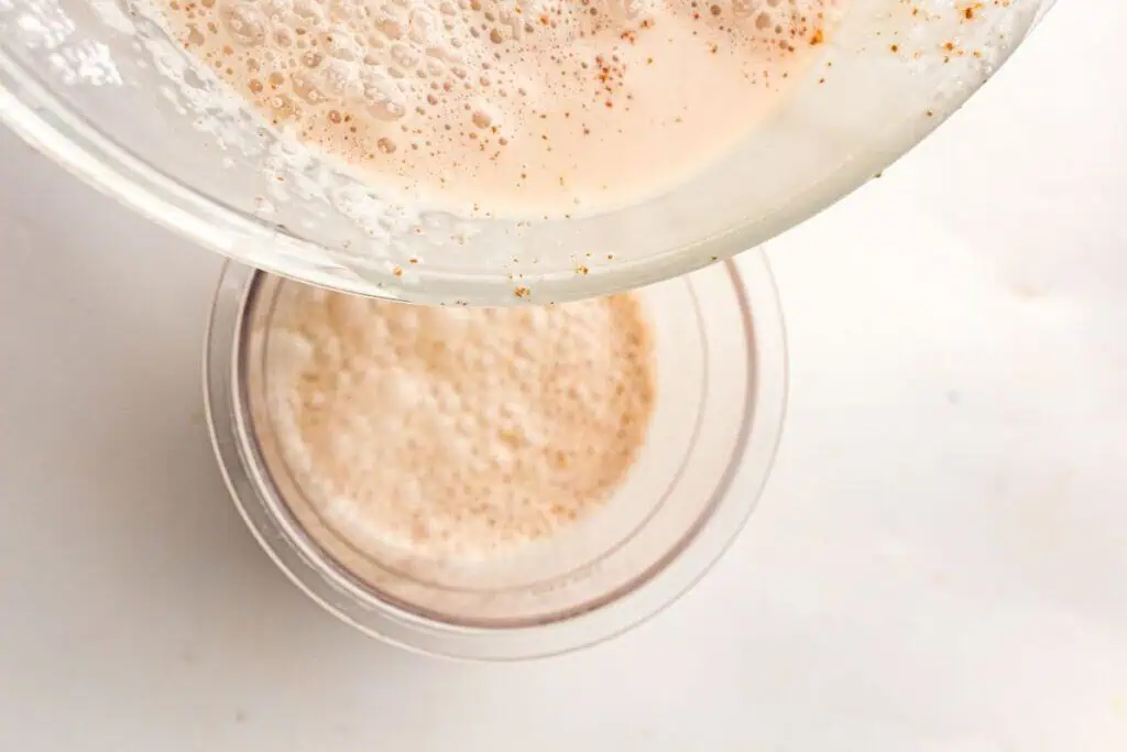A glass bowl with a frothy, light brown liquid is being poured into a clear cup containing a similar foamy beverage, seen from above on a white surface.