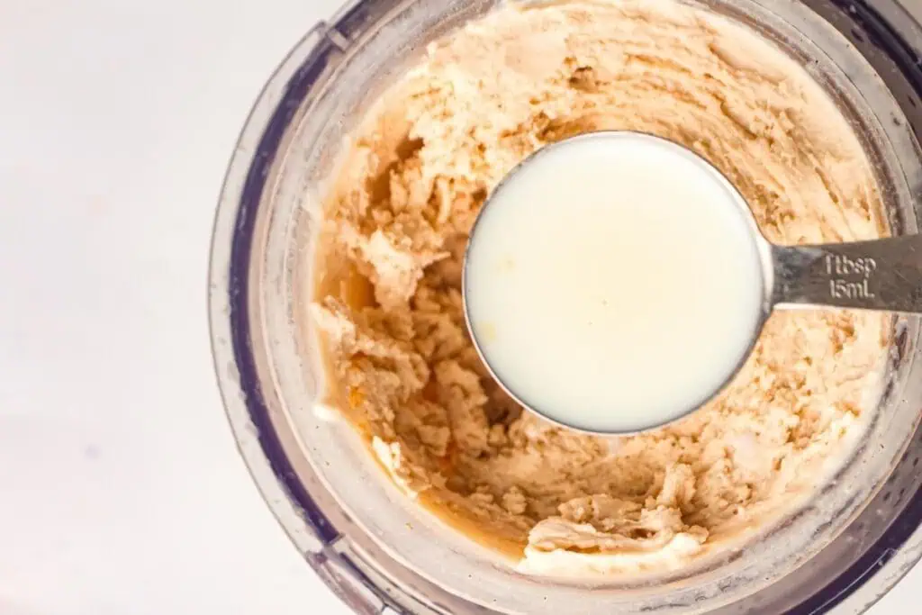 A metal tablespoon filled with milk is held above a mixing bowl containing a creamy, beige mixture.