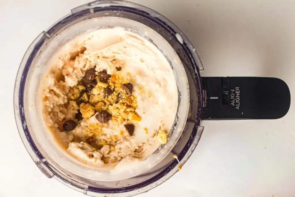 Overhead view of a food processor bowl containing a creamy mixture topped with chocolate chips and crushed cookies.