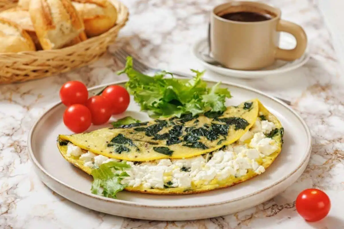 A plate with a spinach and goat cheese omelette, fresh lettuce, and cherry tomatoes, next to a cup of coffee and a basket of bread rolls on a marble table.