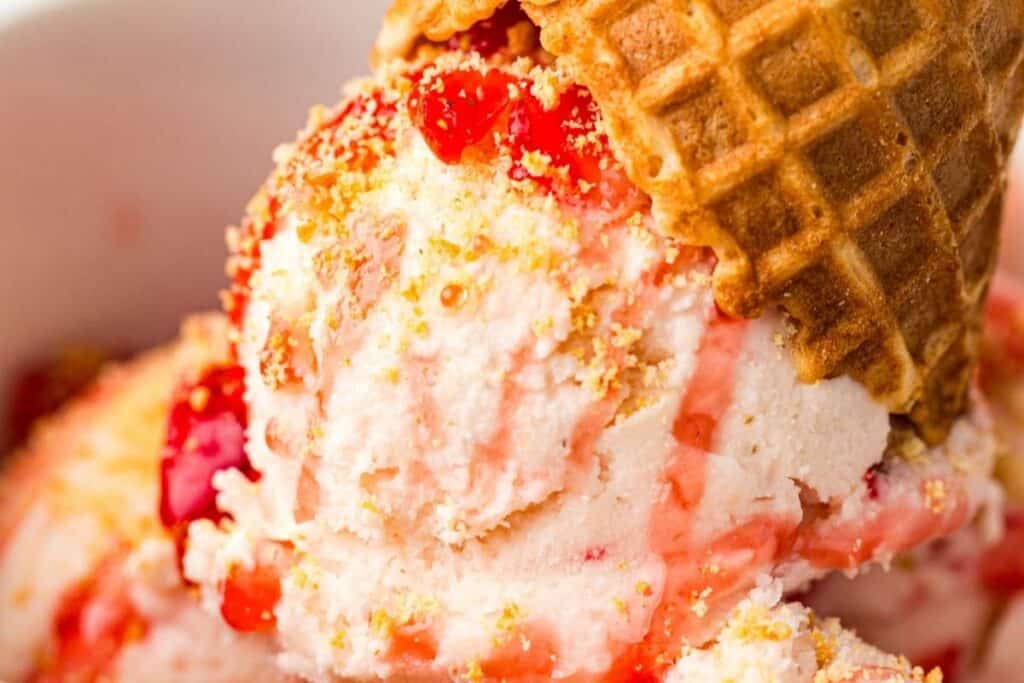 Close-up of scoops of vanilla ice cream with red syrup and crumbs, topped with an upside-down waffle cone.