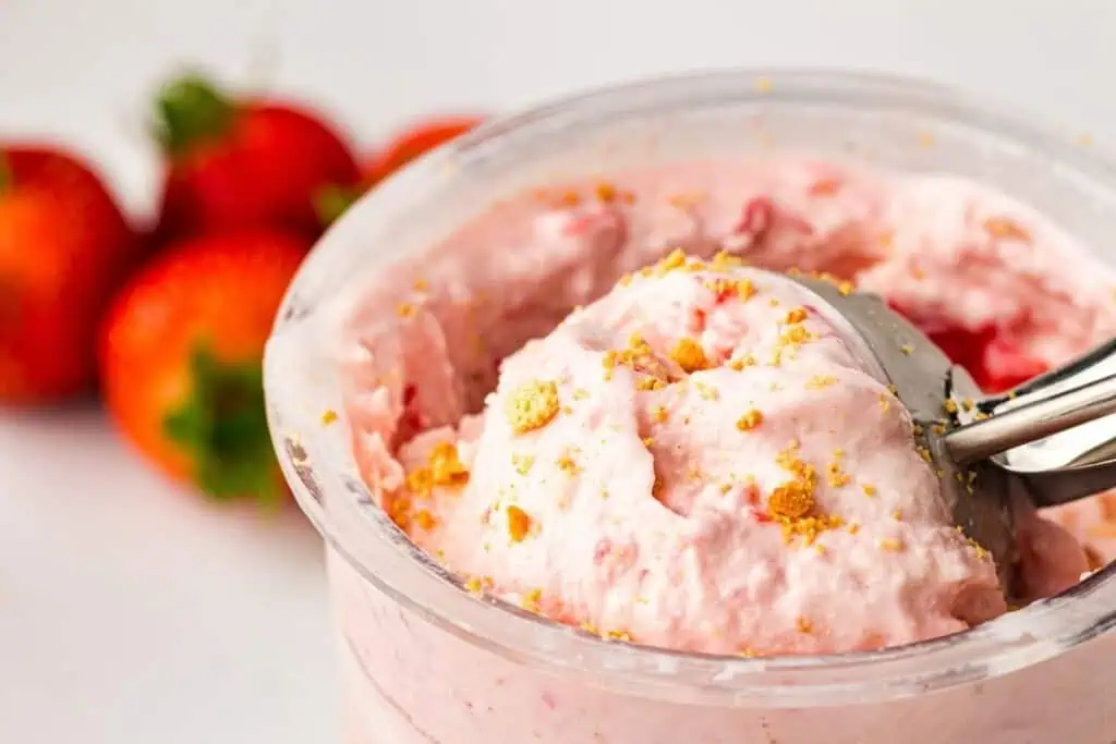 Close-up of a container of strawberry cheesecake ice cream with a metal scoop, garnished with crumbled cookies; fresh strawberries are in the background.