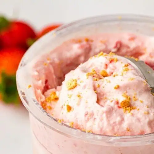Close-up of a container of strawberry cheesecake ice cream with a metal scoop, garnished with crumbled cookies; fresh strawberries are in the background.