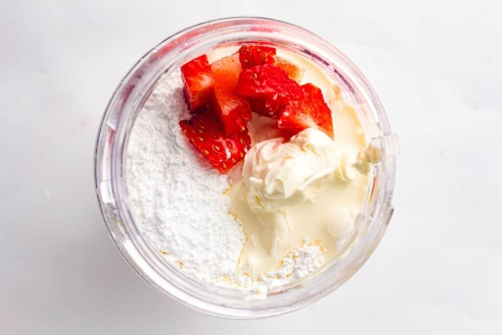 Overhead view of a blender cup containing powdered sugar, chopped strawberries, cream cheese, and condensed milk on a white surface.