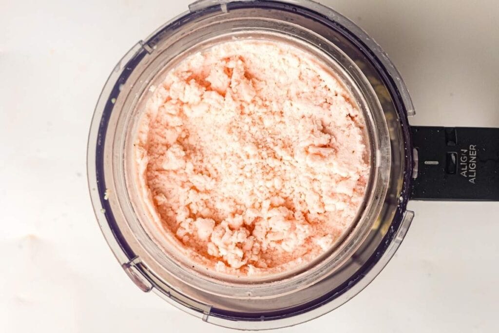 A food processor bowl filled with coarsely blended, pale pink mixture on a light surface.