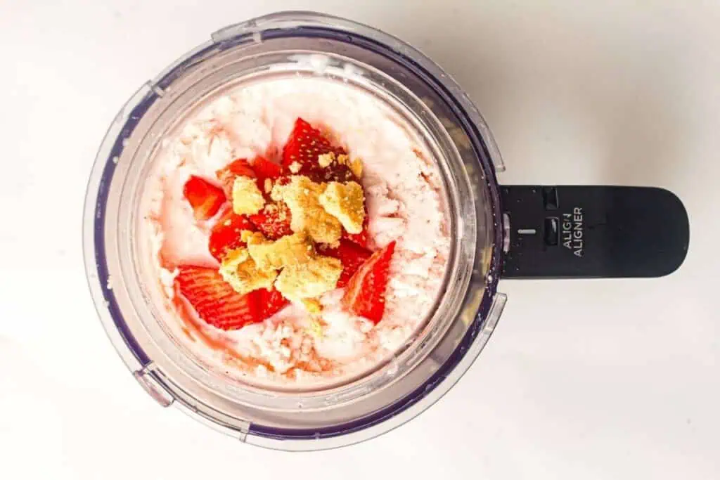A food processor filled with chopped strawberries, chunks of crumbly yellow cake, and a creamy mixture, viewed from above.