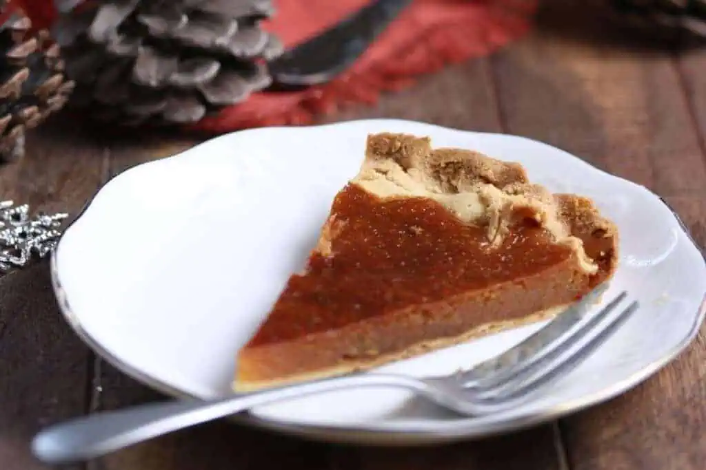 A slice of sugar pie on a white plate with a fork, with pinecones and a red napkin in the background.