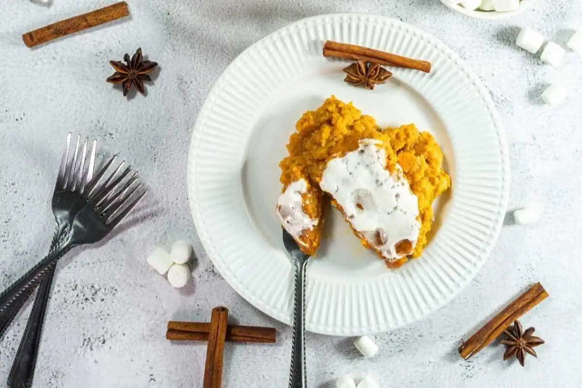 A plate with a serving of sweet potato casserole topped with marshmallows, surrounded by forks, marshmallows, cinnamon sticks, and star anise on a white surface.