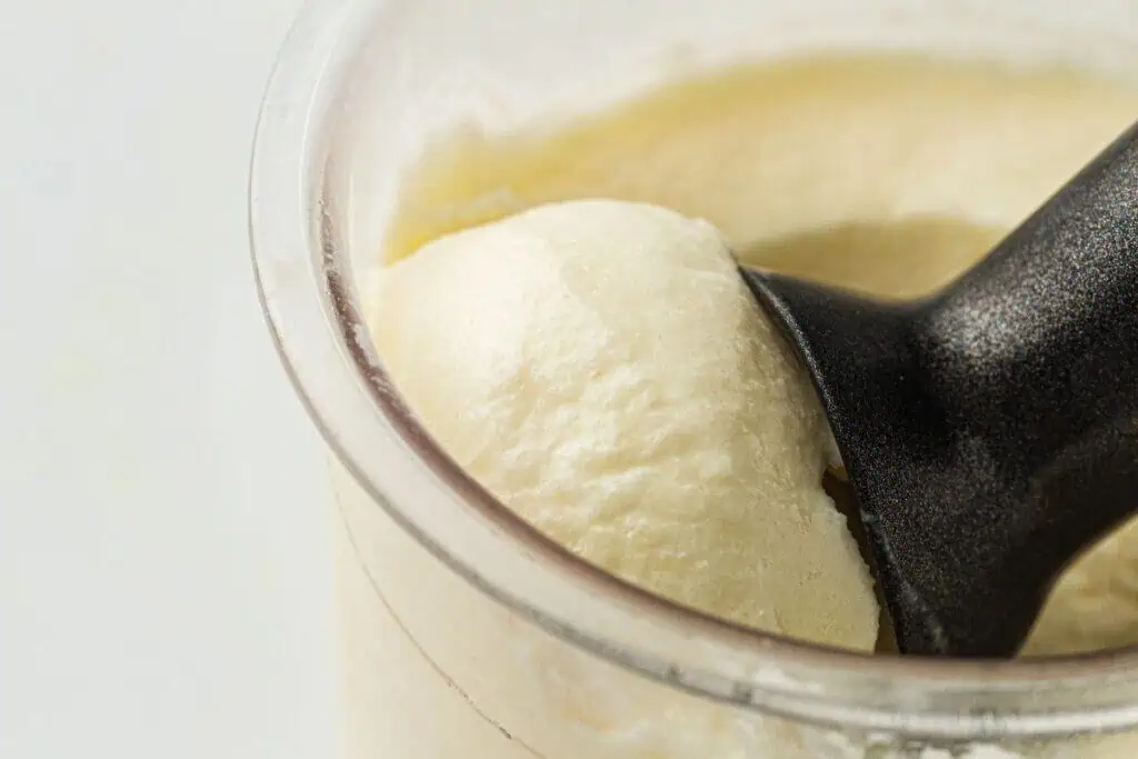 Close-up of vanilla ice cream being scooped from a transparent container with a black ice cream scoop.