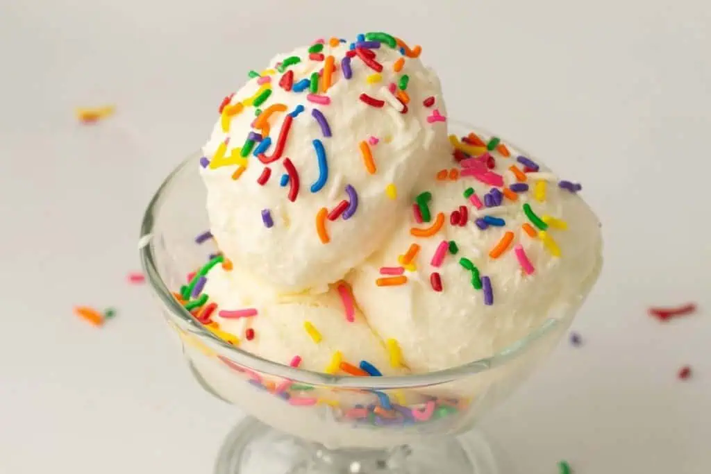 A glass bowl with three scoops of vanilla ice cream topped with colorful rainbow sprinkles on a white background.