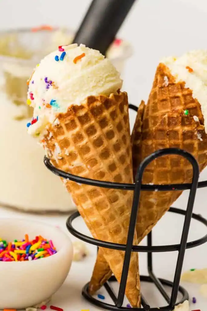 Two waffle cones filled with vanilla ice cream and topped with colorful sprinkles are held in a metal stand; a bowl of sprinkles is nearby.