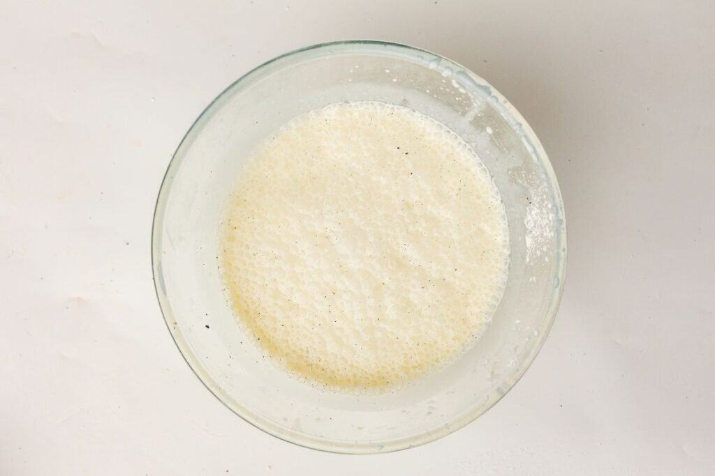 A glass bowl filled with a frothy, creamy liquid mixture, viewed from above on a white surface.
