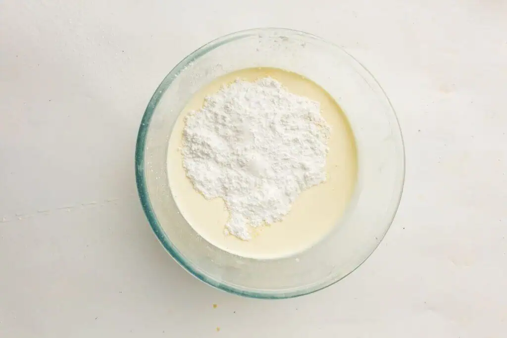 A glass bowl containing cream with a mound of powdered sugar on top, viewed from above on a white surface.
