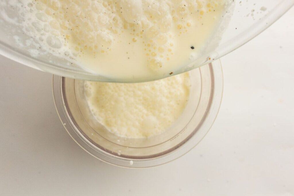 A bowl of frothy liquid, possibly a batter or egg mixture, is being poured into a transparent container on a white surface.