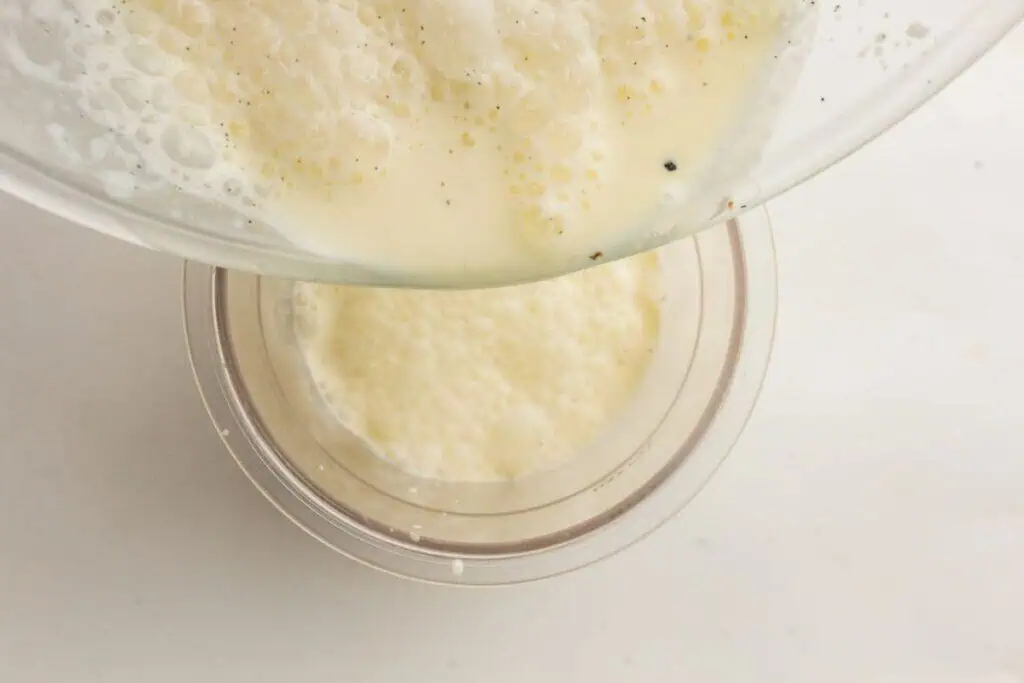 A bowl of frothy liquid, possibly a batter or egg mixture, is being poured into a transparent container on a white surface.