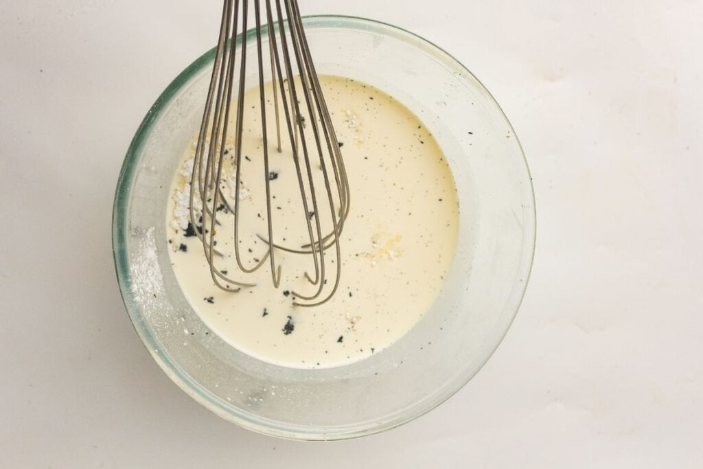 A glass bowl filled with a creamy liquid mixture and black specks, being whisked with a metal whisk on a white surface.