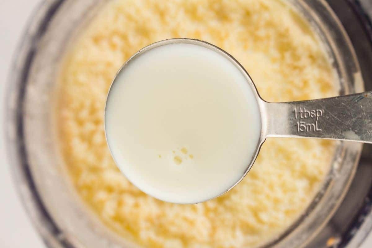 A metal tablespoon filled with liquid buttermilk is held above a bowl containing curds or separated milk solids.