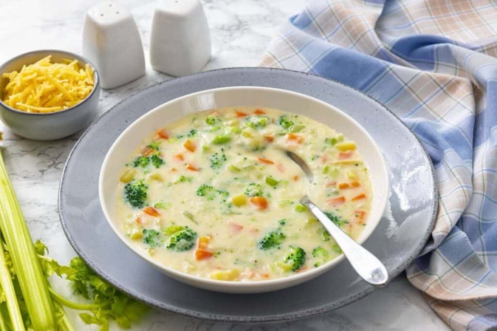 A bowl of creamy vegetable soup with broccoli, carrots, and celery sits on a plate with a spoon, next to a small bowl of shredded cheese and salt and pepper shakers.