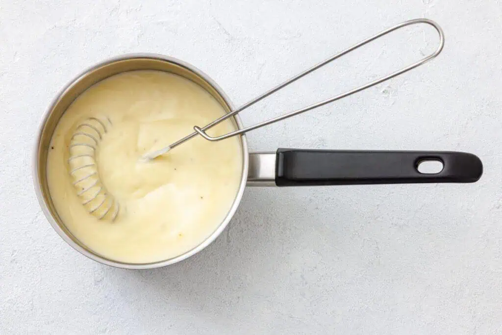 A metal saucepan with a black handle contains creamy white sauce being stirred with a spiral whisk, placed on a light-colored surface.