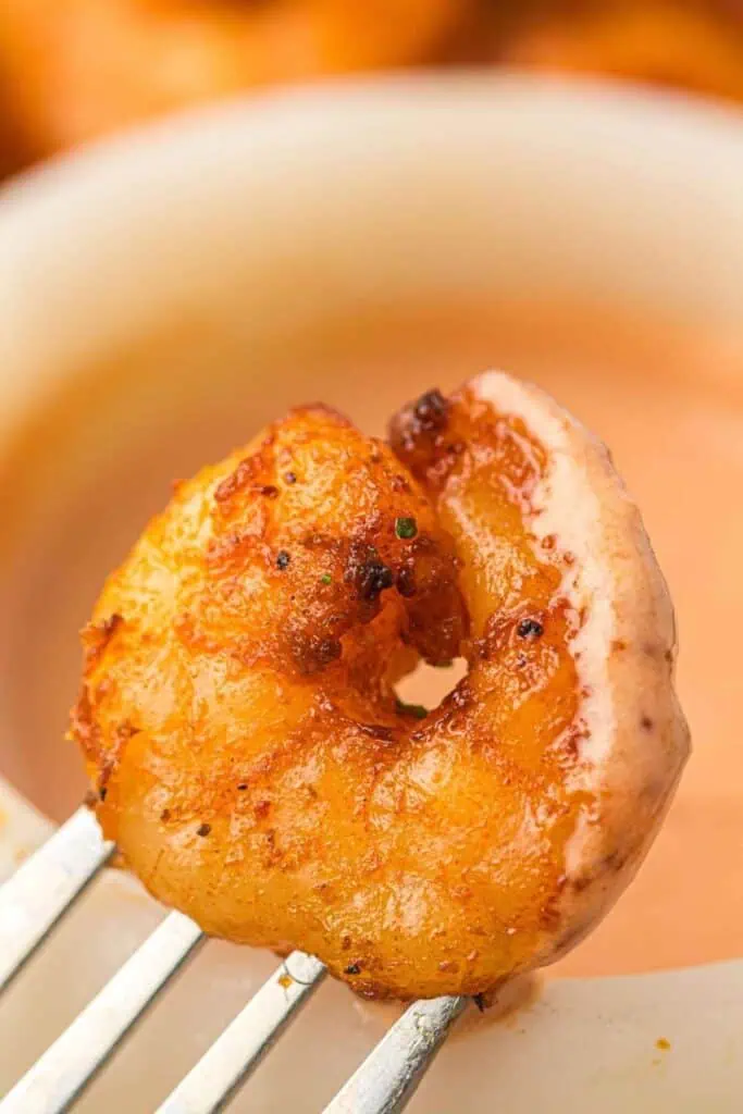 A seasoned, cooked shrimp on a fork is held in front of a bowl of dipping sauce.