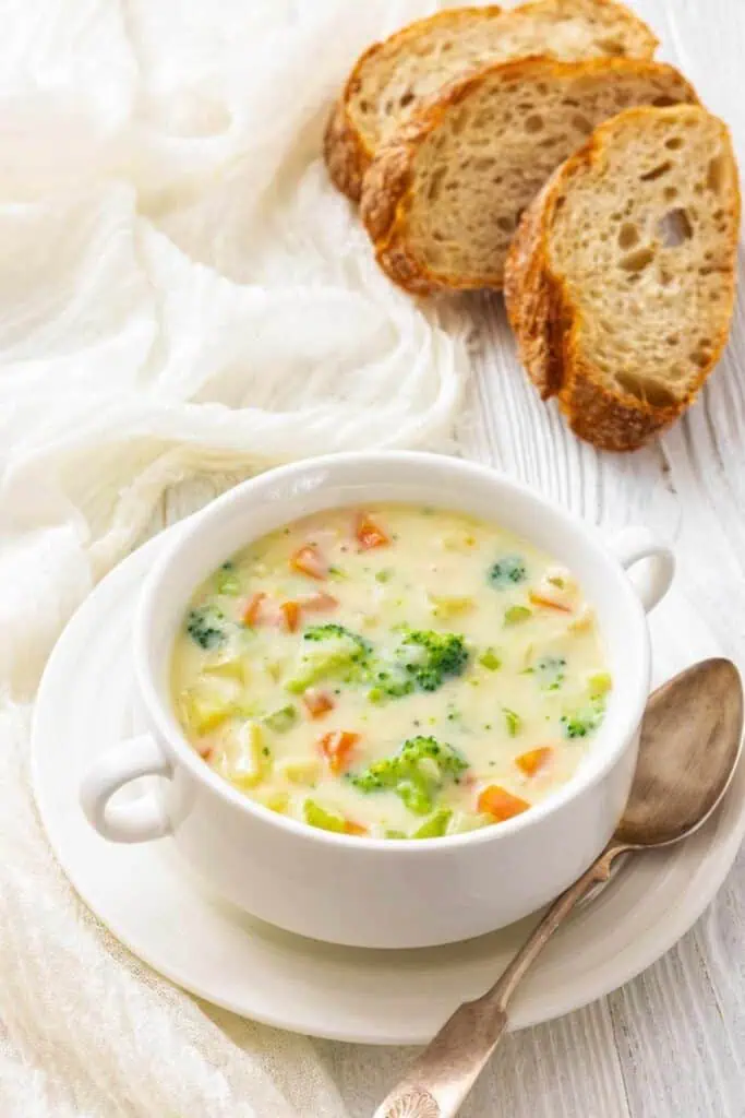 A bowl of creamy vegetable soup with carrots and broccoli, served with slices of rustic bread on a white plate and a spoon.
