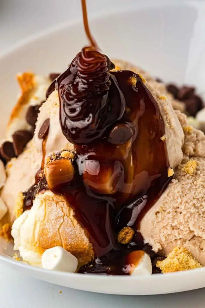 A close-up of ice cream scoops topped with chocolate syrup, marshmallows, and small candy pieces in a white bowl.