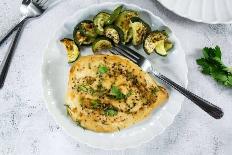 A cooked chicken breast garnished with herbs is served on a white plate with roasted zucchini and a fork.