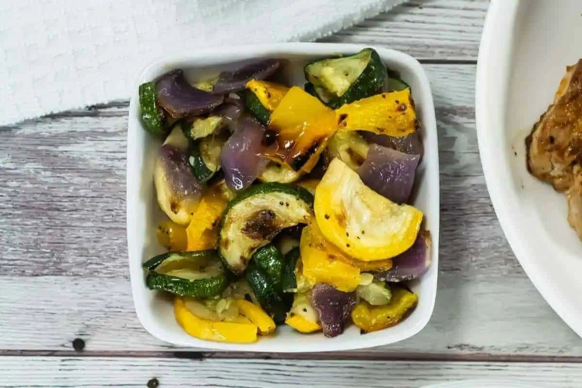 A white bowl filled with broiled vegetables, including zucchini, yellow squash, and red onion, sits on a wooden surface next to a white towel.