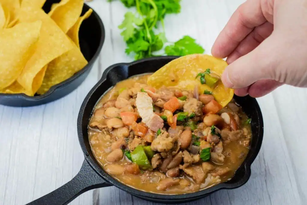 A hand dips a tortilla chip into a small cast iron pan filled with bean and vegetable stew; a bowl of chips and cilantro are in the background.