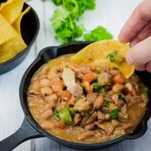 A hand dips a tortilla chip into a small cast-iron skillet filled with Charro Beans With Chorizo, more chips and cilantro are in the background.
