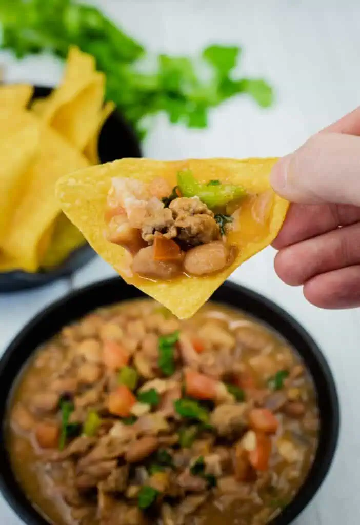 A hand holds a tortilla chip topped with beans, ground meat, and vegetables over a bowl of chili with chips and cilantro in the background.