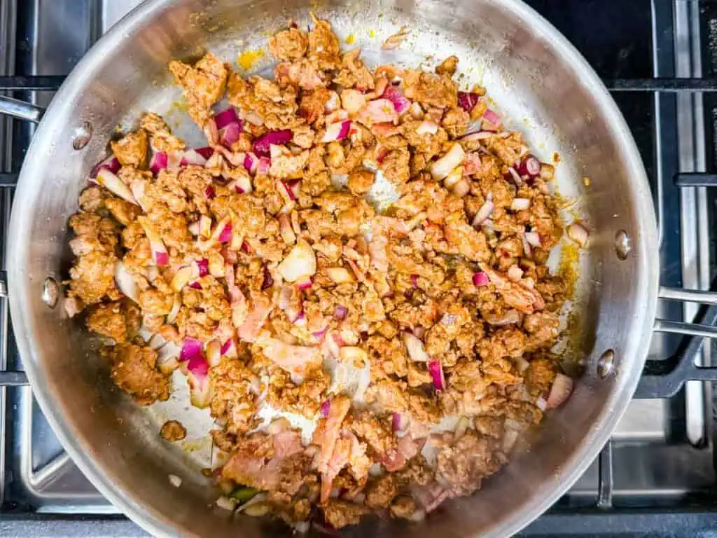 Ground meat, chopped red onions, and bacon pieces cooking in a stainless steel skillet on a stovetop.