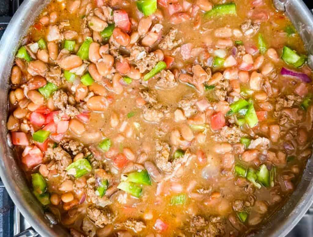 A close-up of a pan filled with chili containing ground beef, pinto beans, chopped tomatoes, green bell peppers, and onions in a thick sauce.