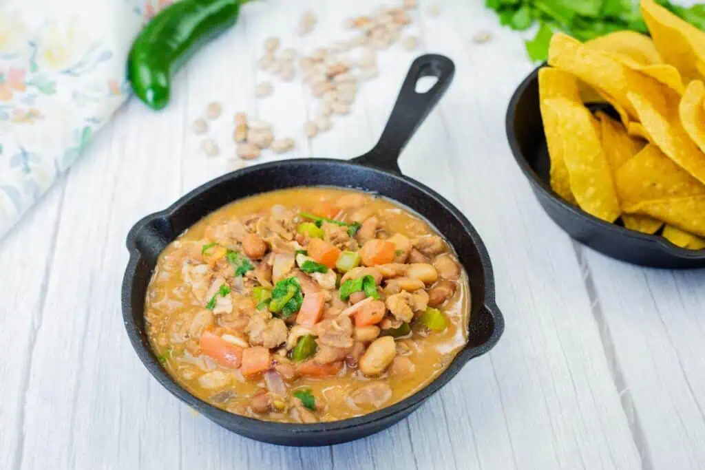 A small cast-iron skillet filled with stew containing beans, vegetables, and herbs sits next to a bowl of tortilla chips on a light wooden surface.