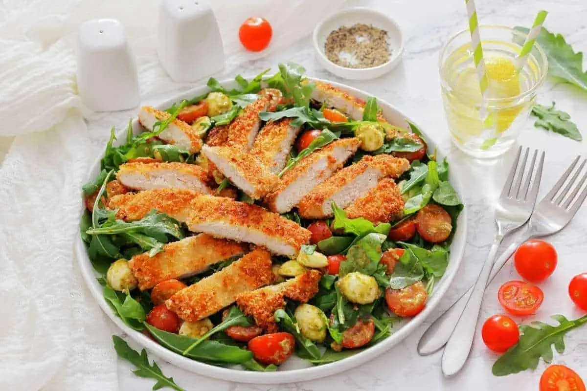 A plate of Chicken Parmesan Salad with a glass of lemon water and utensils on the side.