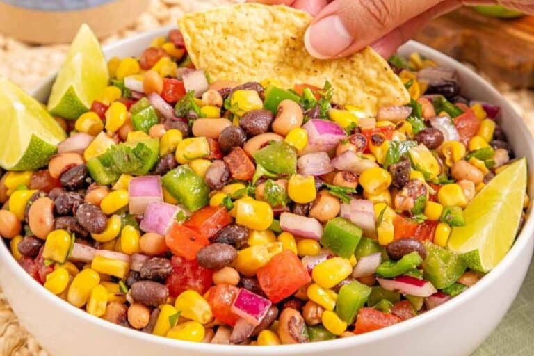A bowl of Cowboy Caviar, garnished with lime wedges; a hand dips a tortilla chip into the salad.