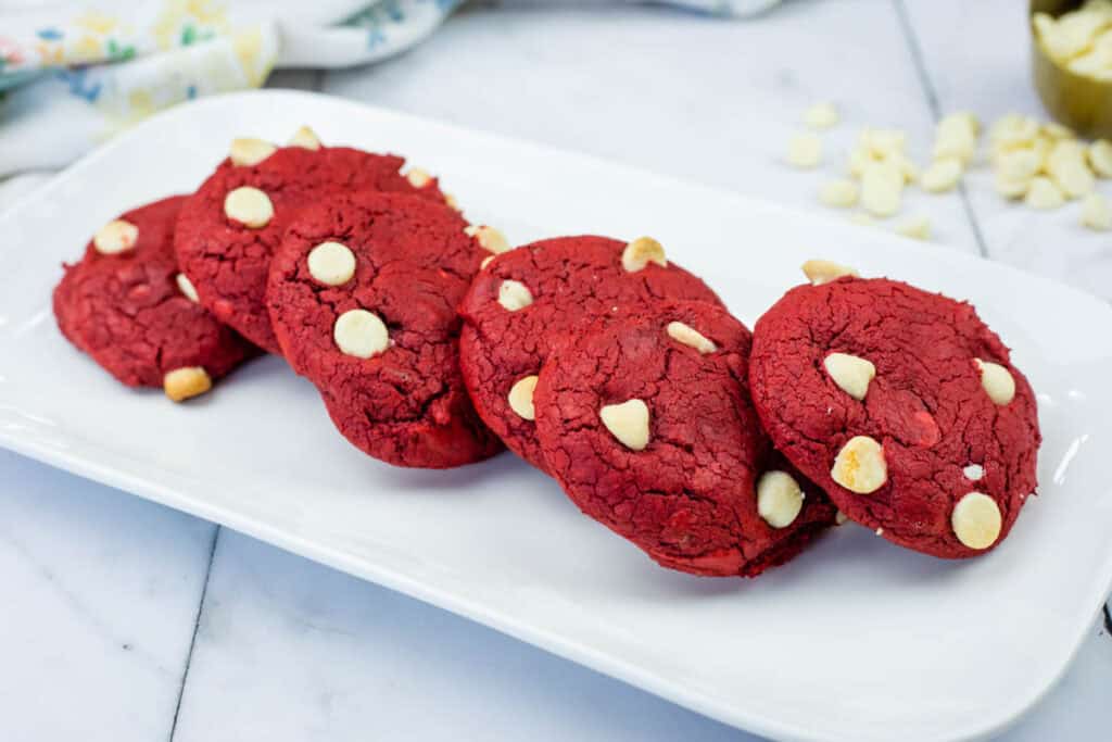 Five red cookies with white chocolate chips are arranged in a row on a rectangular white plate, set on a marble countertop.