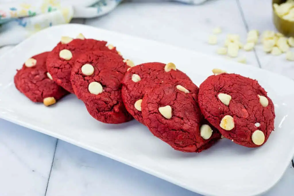 Five red cookies with white chocolate chips are arranged in a row on a rectangular white plate, set on a marble countertop.