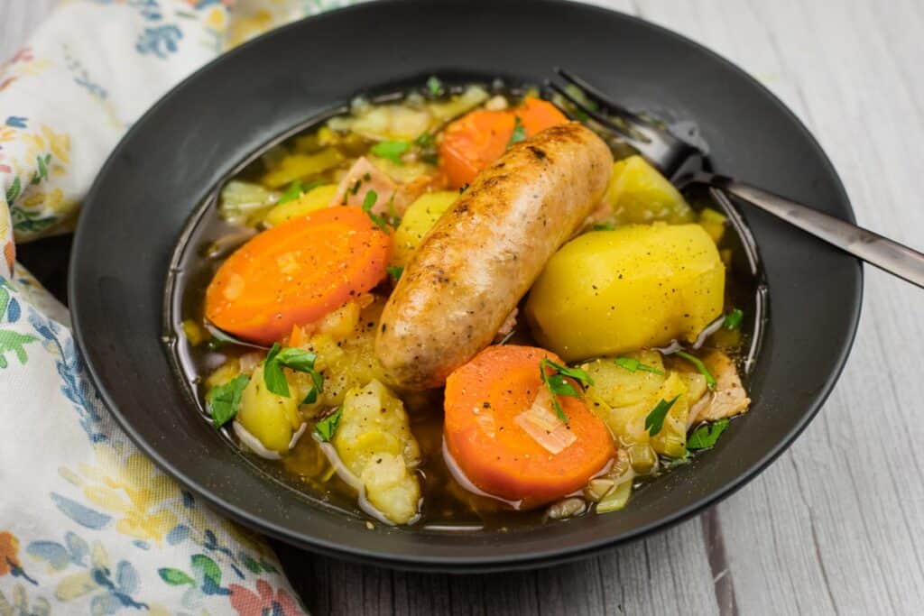 A black bowl filled with Dublin Coddle containing potatoes, carrots, celery, and a sausage, garnished with herbs, with a fork resting on the side.